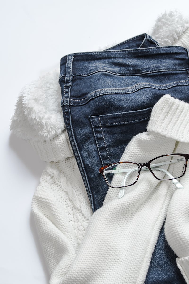 Stylish flat lay featuring denim jeans, white sweater, and eyeglasses on a bright background.